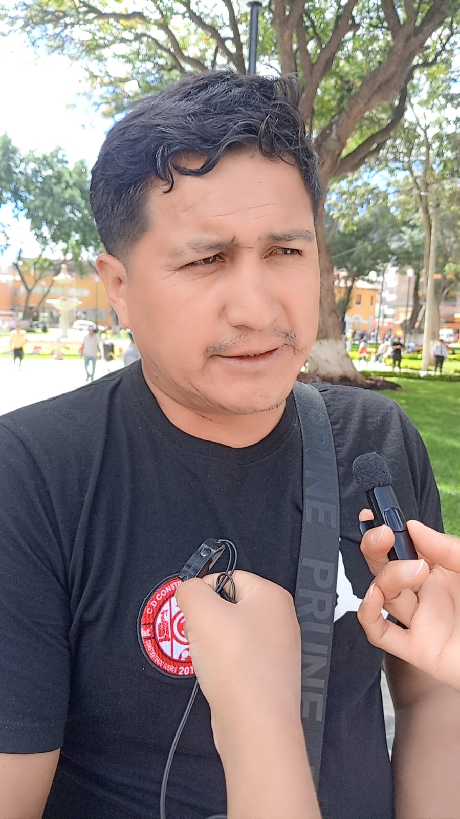 Un hombre con camiseta negra es entrevistado en un parque. Un micrófono de solapa está conectado a su camiseta.