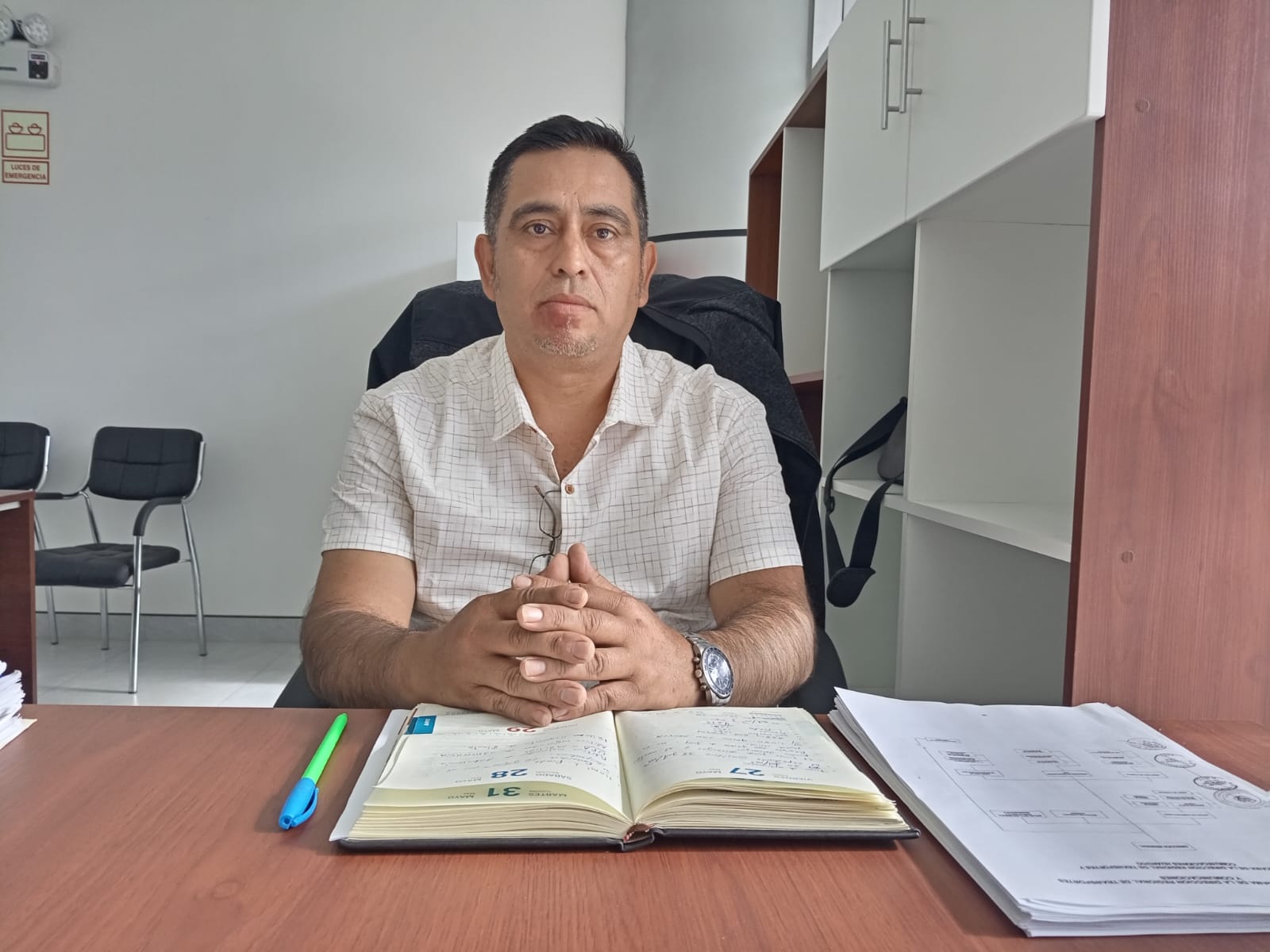 Un hombre está sentado en un escritorio con un cuaderno abierto y documentos. Detrás de él hay una estantería.