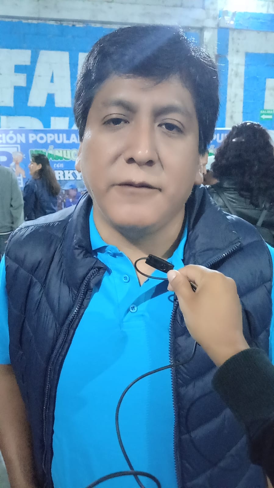 Primer plano de un hombre con camisa azul y chaleco, sosteniendo un micrófono de solapa. Al fondo, un letrero azul con texto.