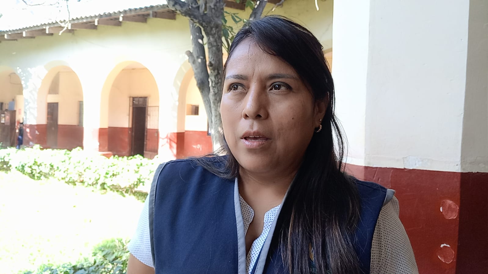 Una mujer morena con el pelo largo y oscuro mira hacia un lado. Detrás, un edificio con arcos y vegetación.